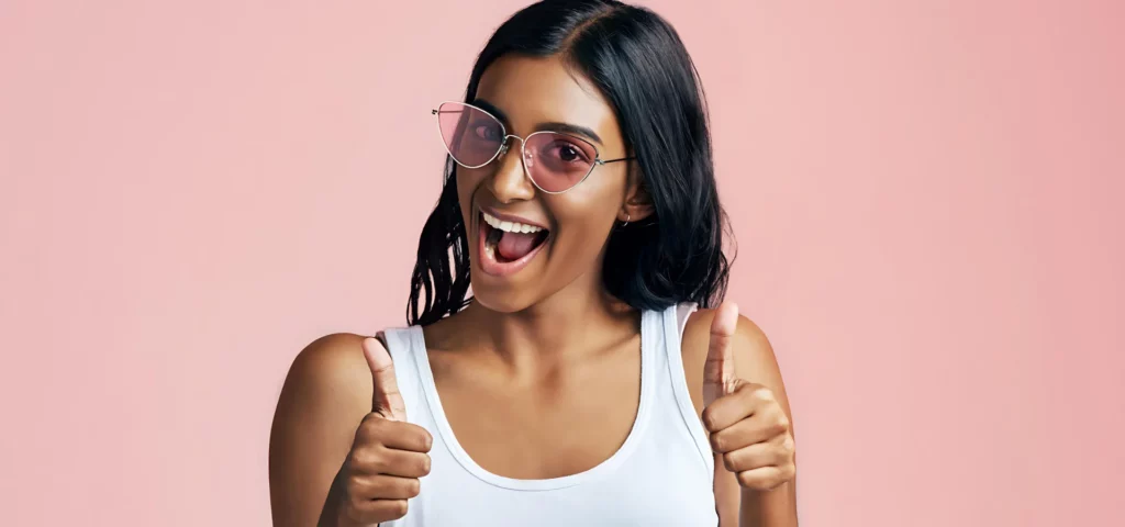 a young woman is in the center of the image. she is wearing a white strapless shirt. and pink sunglasses. she has long black hair and a darker skin tone. she is holding her thumbs up and her smile is wide with her mouth slighlty open. she is on a pink background.