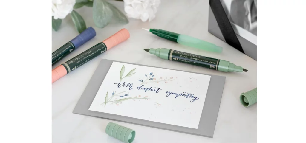 in the center of the image there is a card that has been made. it is a white card sitting on a silver envelope. the card says with deepest sympathy is cursive. there are three watercolour markers open around it showing theor double sided nibs. they are green, blue and pink markers open and a watercolour brush open behind them. they are resting on a table. the markers are sold by my art shop