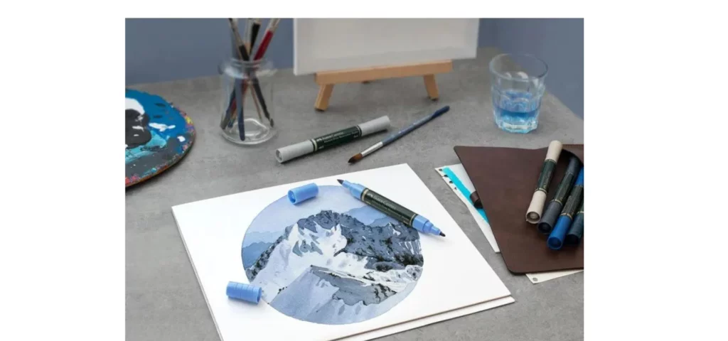 in the center of the image is a round drawing of snow covered mountains in shades of blue, grey and white. they are resting on a table next to a varity of markers that are both open and closed. they are sitting infront of a jar with water and an easel.all sold at my art shop