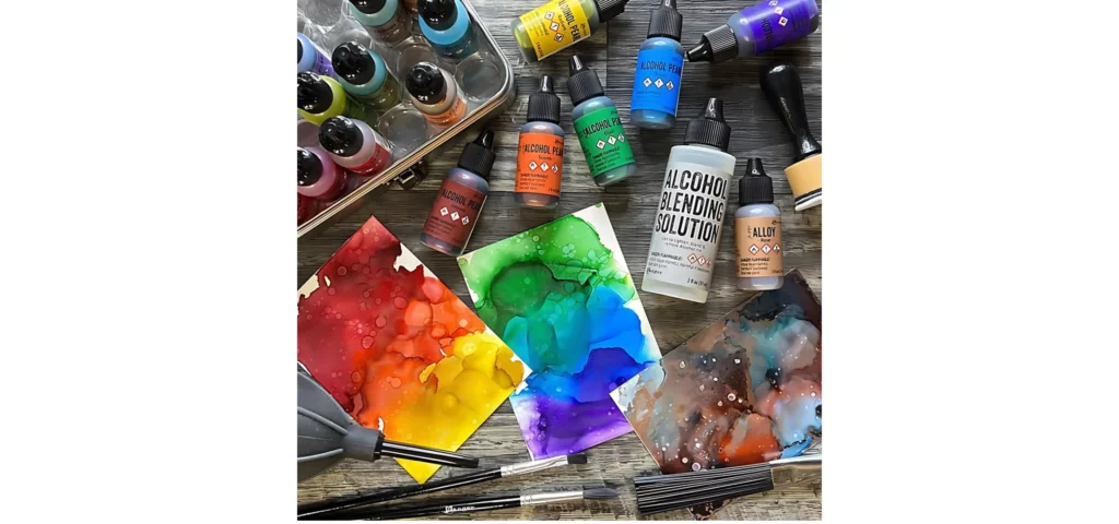 in this image there are many different alcohol inks layed out on the table and some in the tin to the top left of the image. in the center are two card stocks of alocohol ink backgrounds in a rainbow colour. vibrant reds, yelows and oranges and the other blue and green. infront of them are the tool sets which are the plastic brushes. they are resting on a wooden tabletop. its a bright happy image