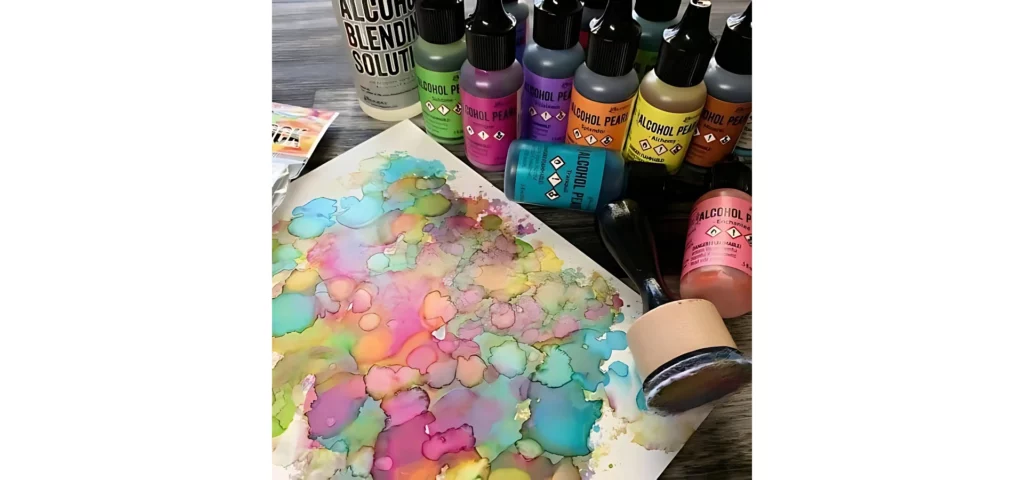 in this image. to the right hand side there are various bottles of alcohol ink standing up and two lying down. they are all in different colours. next to them toawrds the bottom right of the image is an applicator tool that is lying down. they are resting on a yupo paper that has been inked using more pastel pearl tones in pinks, blue and green. the light is coming from the bottom left and casting shadows on the right of the products. they are resting on a wooden tabletop.