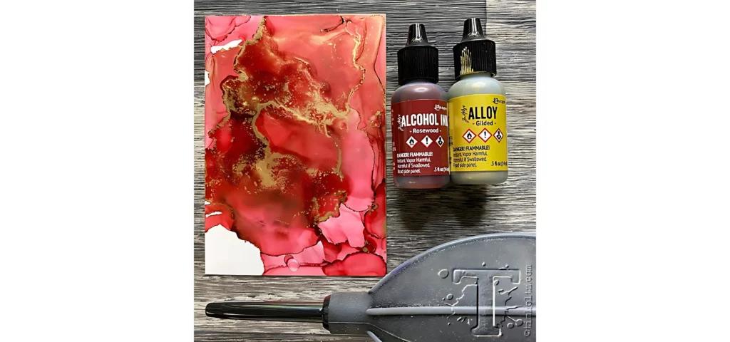 alcohol-ink-alloys-crafting-wizardry. in the center of the image there are two alcohol ink bottles lying down and name facing forwards. they are a red and an alloy. to the left of them is yupo paper that has been inked on using those two inks on the right of it. the yupo is shades of red and has gold details on it. below it is an alcohol ink blower lying on its side. they are all resting on a dark wooden tabletop.