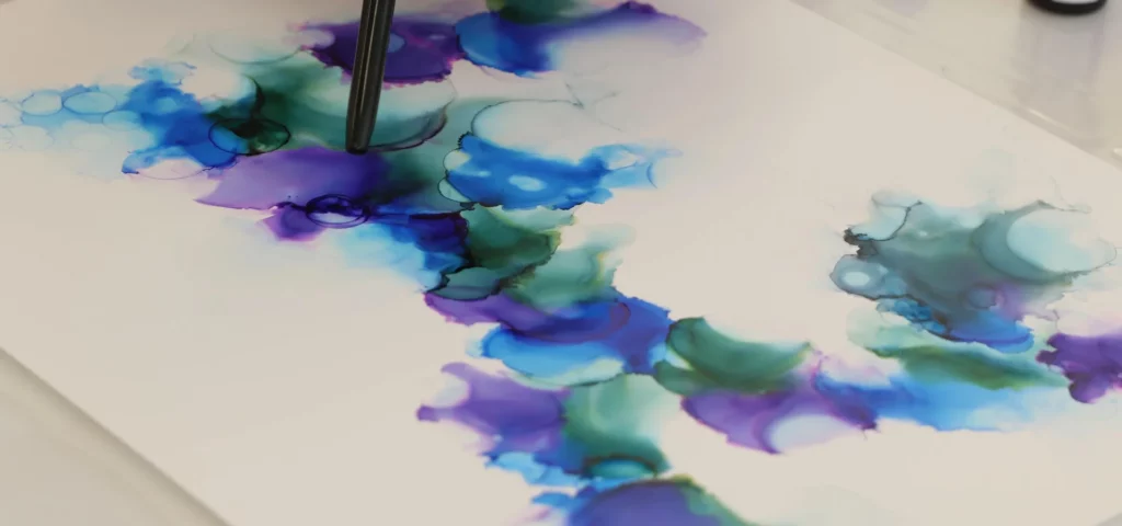 in this image in the center there is a yupo paper that is slightly off center. on it are the inks in colours of blue and green and purple. they are all blending together. there is a nozzle touching the paper at the top left hand side on the ink colour. in the top right corner is another bottle of alcohol ink. on a white surface.