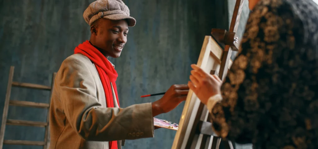 in this image there is a man on the left hand side painting agaisnt an easel. he is wearing a jacket, red scarf and a hat. holding a paintbrush. there is a woman on the right hand side of the image standing behind the easel and canvas. behind the man is a dark green wall and a ladder. the light is coming from the top right hand side of the image. the man is smiling and talking while he paints.