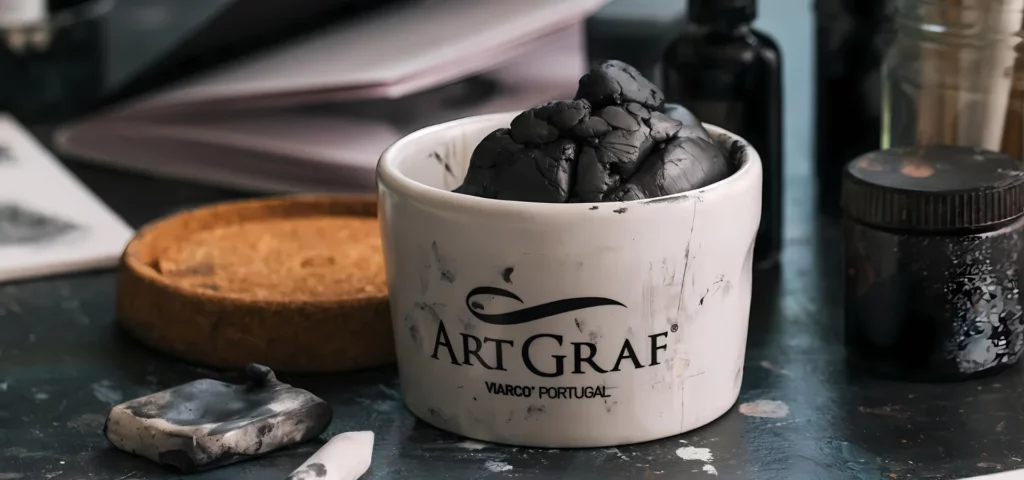 in the center of the image is a pot with the name artgraf writen on it in black. inside the pot is a ball of used and disfigured graphite putty. the lid is cork and sitting to the left of the open jar. around the jar are various other art tools. on a dark work surface, with a book in the background. 