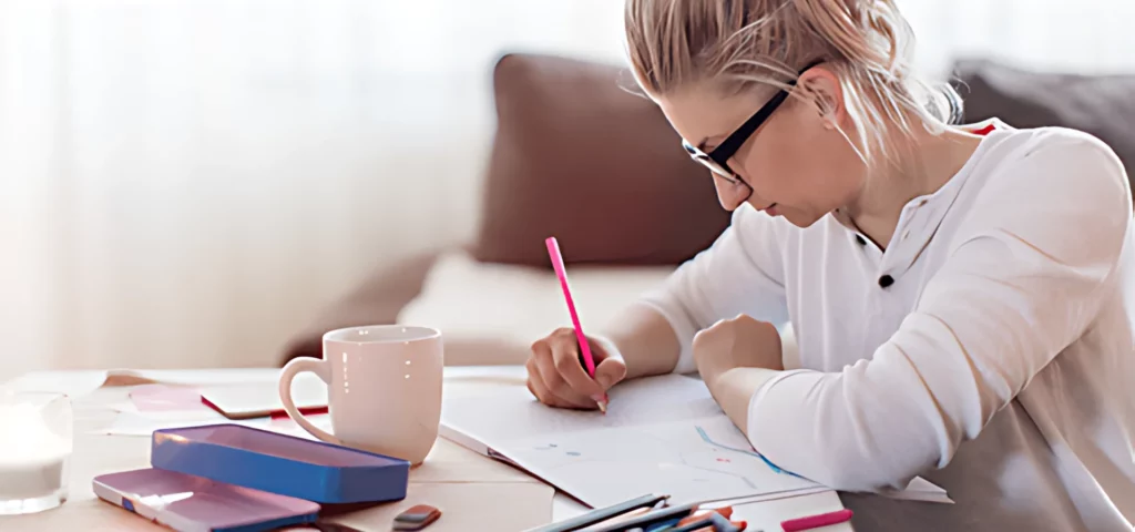 to the right of the image is a woman wearing a long sleeve white shirt sitting down and leaning over a desk and colouring in using a pink pencil. she is leaning on the table with her left arm. there are a stakc of papers beneath her. infront of her is a cup of coffee and a tin of coloured pencils. the background is a blurred image of a lounge as you can see a brown leather couch in the background.