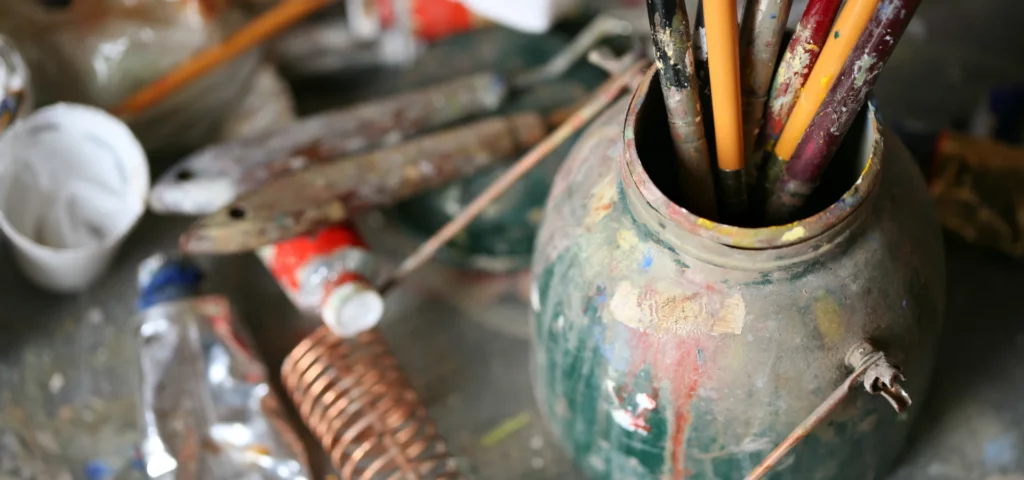 in this image there is a pot of brushes that need brush cleaning. they are sitting to the right hand side of the image in a very dirty glass jar that is full of paint stains and next to it on the left are various tubes of paint and palette knives that are also covered in used paint and are looking dirty and in need of a clean. they are resting on a table that has dried paint on it. the light is coming from the left side of the image. the colours in the image are mainly blue, green, yellow and red tones.