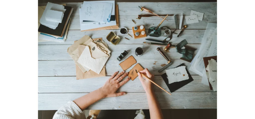 in the center of the image there is a table with a collection of inks and pens scattered over it. there is a person sitting st the bottom of the image using a quill on a brown paper. the table is a light white wooden table. there are books towards the top of the table.