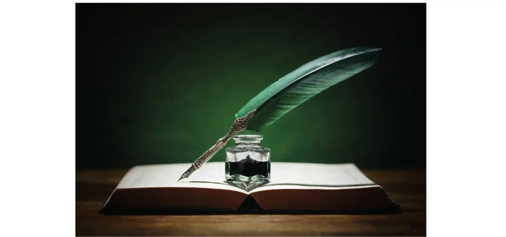 in the center of the image there is an open thick book. in the center of the book is a clear glass jar that has black ink in it. resting on it is a quill with a deep green feather. the background is a deep green wall. on a dark brown table. pens sold at my art shop