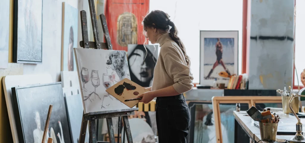 there is a woman in the center of the image painting on a canvas. she is wearing a beige shirt and black pants. she is holding a square wooden palette that has paint on it in her left hand. her right hand is not visable. she is painting a large canvas that is a rectangle shape and that has pictures of wine glasses on it. it is standing on an old paint covered easle towards the left of the image. all around her are canvas in various sizes that have been painted on. behind her is a table with paints and other art supplies on it. the light is coming from behind her so she is a little framed by the light.