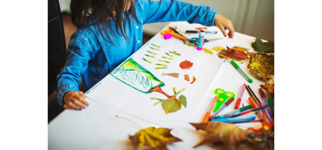 on this image there is a child dressed in blue sitting on the left hand side with a table infront of them. on the table are a variety of pens and pencils for colouring in. and paper that has been drawn on. htere are dried leaves scattered across the table, the light is coming from infront of the child from the right hand side of the image.