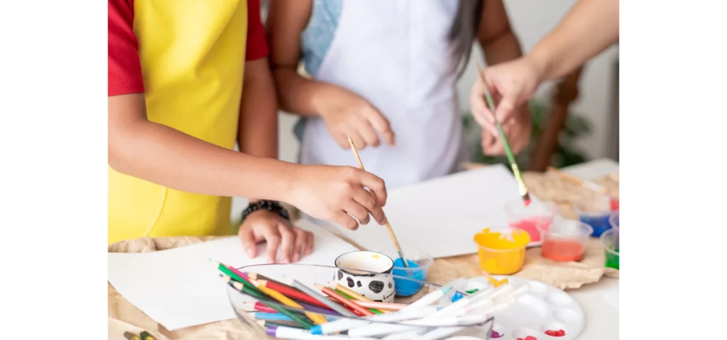 in this image there are two children in the image wearing aprons and holding paintbrushes and painting on the paper on the table infront of them. they are painting in blue and yellow colours. next to where they are painting there are pencils and crayons lying on the paper.