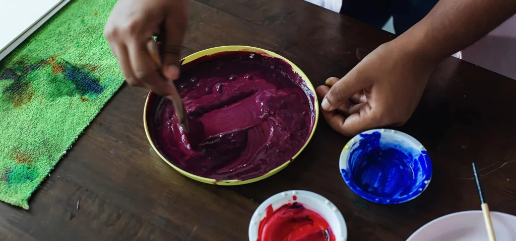 in the center of the image there are three bowls on the brown wooden table. there are two white small bowls on the right with red and blue paint in them and on the left is a larger yellow bowl with a purple in it. there is a person standing in the top right of the image holding a wooden spoon stirring the purple paint. their stirring hand is slighlty blurred as its an action shot. there is a green micr fibre cloth on the left of the bowls that has paint on it from being used to wipe hands. the light is coming from the left of the image so the shadows are casting to the right of all the bowls.