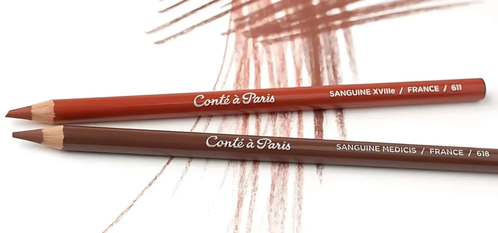 two conte pencils are sitting in the center of the image. they are lying with their tips facing the left side of the image. they are resting on a paper that has been coloured in using those pencils in the picture. they are both a red brown colour and have their name clearly written on it in a silver. 