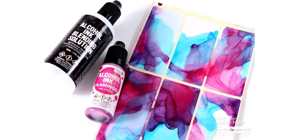 the couture alcohol inks are shown lying down in the image on the left hand side. next to a bottle of blending solution from the same brand. the bottle has a white label with a colour circle of what colour is inside and the blending solution bottle has a black label with the name in white. both have black lids. they are lying next to a yupo paper that has been inked with blue and pink. on a white background