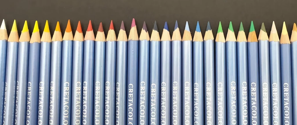 Cretacolor Marino Watercolor Pencils Elevate your Artistry shows the pencils lined up acrosee the bottom if the image. they are facing their nibs upwards to the top of the image. they have metallic blue wraps around each pencil and the names of the pencil is clearly visable on each one. they are in a rainbow order. from yellows on the left to the browns on the right hand side. they are on a black background