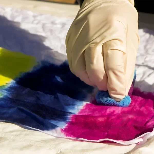 A persons hand is seen coming in from the top of the frame. They have a glove on, and are applying Dala All-In-One Fabric Dye to a piece of cotton fabric. They have applied 3 colours, yellow, blue and pink.