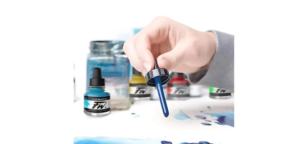 A promotional image for Daler Rowney FW Acrylic Inks. A close up of a persons hand holding the eyedropper from a blue ink and dropping ink onto a piece of paper. There are other bottles of ink in the background that are blurred. The image is cut off by the frame.