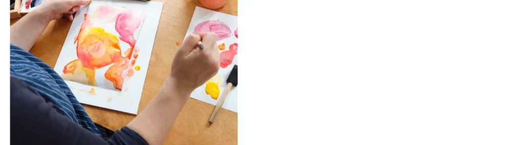 in the center of the image is a woman in blue painting a watercolour painting on apaper and a flat wooden board. she has two sheets of paper infront of her both have the colours of pink and yellow and orange across it. they are blending into eachother. she is holding a brush in her right hand over the oainting.