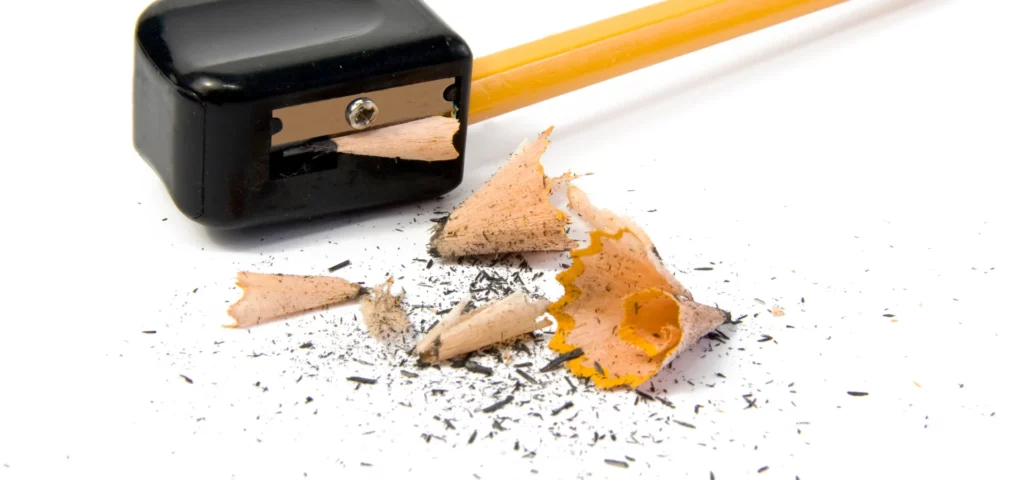 in the center of the image there is a sharpener that is black with the silver metal blade in the action of sharpening a pencil. the pencil is coming from the right hand side of the image and is yellow. in front of the sharpener are the sharpenings from the pencils. bits of wood and graphite laying on the white table. on a white background