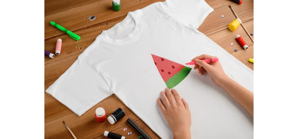 in the center of the image there is an open childs white tshirt. there is a picture of a watermelon on it and someone is holding a pen and colouring it in. there are paints and pens scattered all across the brown tabletop surface.