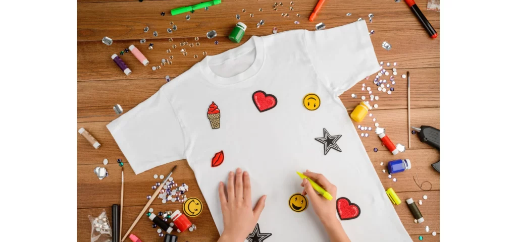 in the center of the image there is a white childs tshirt that has hearts and stars and smiley faces drawn on it. there is a pair of hands from the bottom holding a yellow marker drawing on the shirt. there are various markers and paints scattered across the tabletop surface. the table is a reddish brown wooden table.