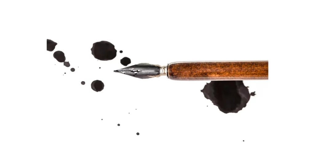 in the center of the image is a brown stemmed calligraphy pen that has its nib facing towards the left hand side. around it are drops of black ink in various sizes. on a white background