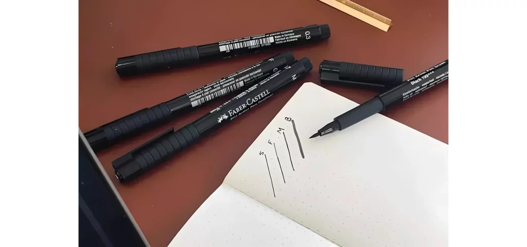 in the center of the image is an open note book that has four lines of different sizes drawn in it. the pens are above the book on a dark wooden table, there is one pen laying on the book. all the pens are black.