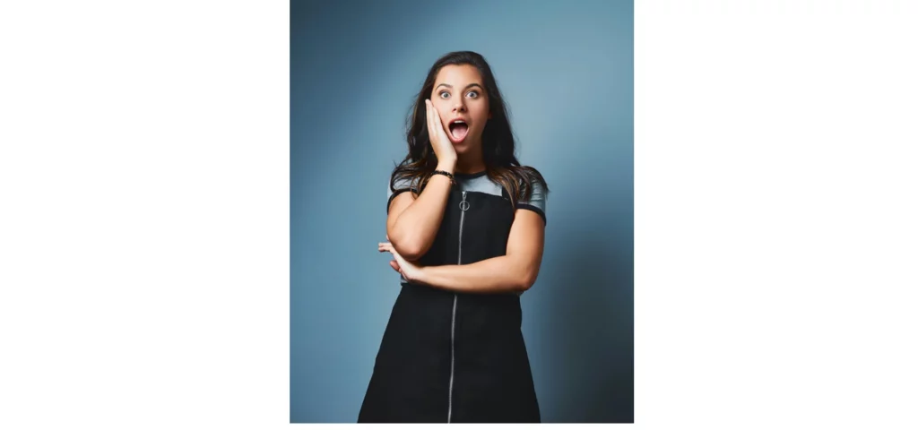 in the center of the image there is a lady standing in a black dress with her right arm across her stomach meeting her left arm which is going up toawrds her face. her hand is on her face and she has a look of suprise on her face. her mouth is open and her eyes are largge. she has long brown hair.on a blue tones backdrop