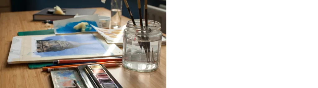 in the center of the image there is a watercolour painting on a table with various pencils and paints around it. infront of it is a glass of water with brushes resting in it. they are all sitting on a table with notebooks and tissues scattered around in the background