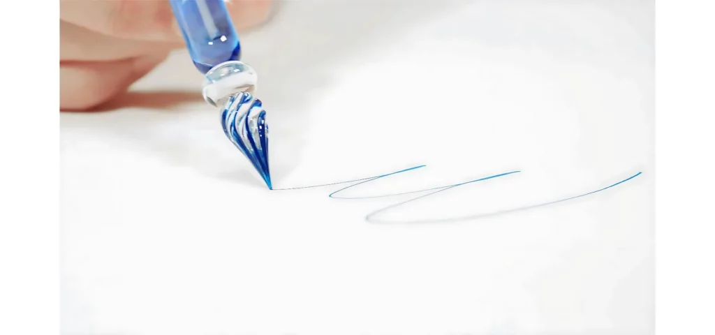 in the center of the image there is a blue glass pen that is being used to write a word in blue ink. the ink is on the white paper. the hand is visable at the top left corner.