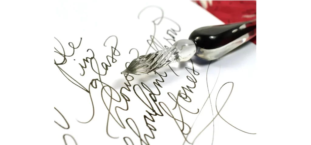 there is a black and clear glass dipping pen in the center of the image. sitting on white paper that has calligraphy writing on it in black ink. behind it is a red colour.