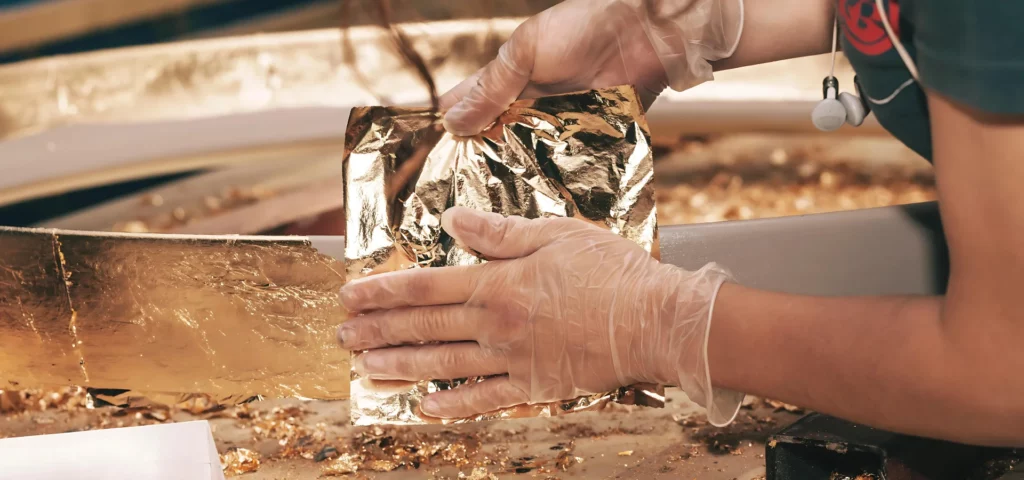 in the center of the image there is a person who is wearing clear gloves and holding gold leah sheets applying them to an object in the center of the image. they are working on a wooden table and there is gold leaf broken off and lying around the table. 