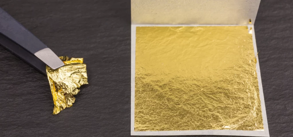 on the right hand side of the image is a gold leaf booklet showing a sheet of gold leaf. it is square and brilliant bright gold. to the left of it is a pair of black tweezers with silver tips holding onto a chunk of the gold leaf. they are both resting on a dark wooden tabletop.