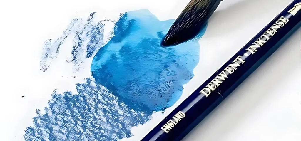 There is a single blue Derwent Inktense Colour Pencil shown laying on a piece of white paper with a sketch swatch on the paper. There is a paint brush that is being run through the swatch to show that the pencil is water soluble. A close up and is cut off by the frame.