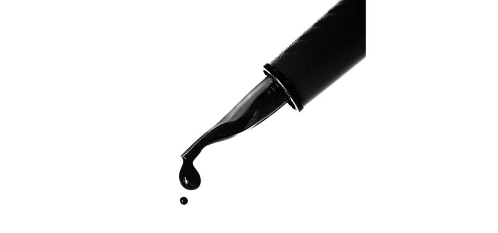 isomars acrylic ink is shown in the image with a black pen coming in from the right hand side it nib is facing towards the bottom of the image. from the very tip of the nib there is a drip of pure black ink falling off of it. on a white background