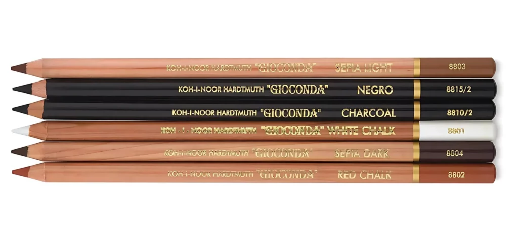 koh-i-noor sketching pencils are spread out in a line in the center of the image. they are lying horizontally on the image with their nibs pointing leftwards. they are a mixture of brown cased and black cased pencils with gold writing on them. they are on a white background