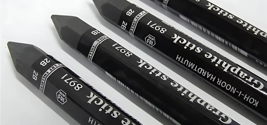 the koh-i-noor graphite sticks are showing as a close up in the center of the image. they are a dark grey colour with a black wrapper around them that has white writing on it describing what each product is. they are nib facing up towards the top left hand corner of the image. they are lying on a white background and the light is coming from directly above as there is a slight shadow casting down on them