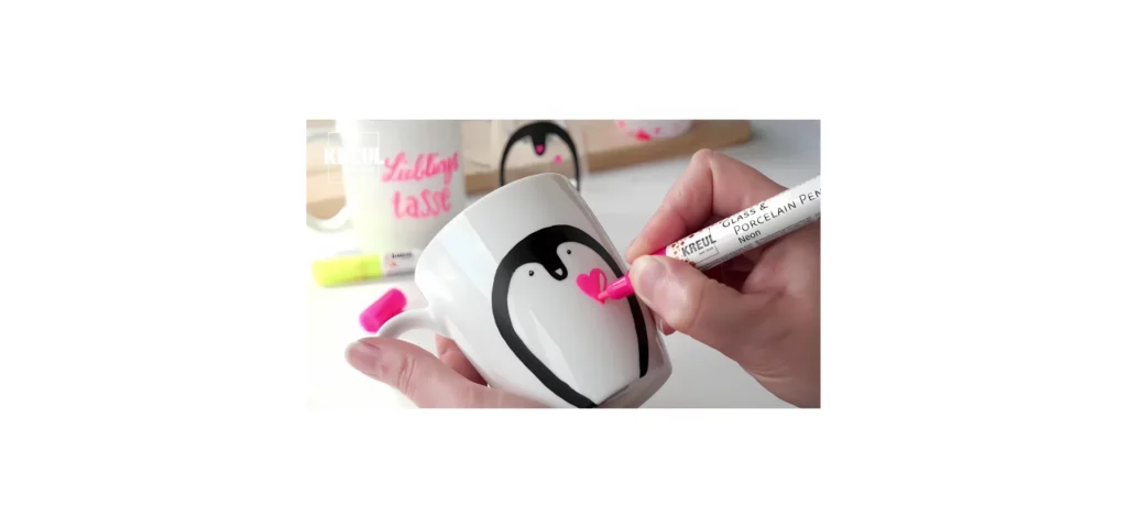 in the center of the image there is a cup being held by a person they are drawing on it with a pink pen. there is an image of a pengiun being drawn on it. the background you can see a varieyt of other cups