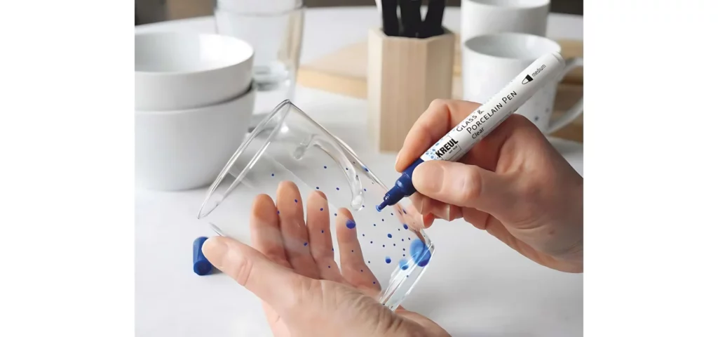 in the center of the image there is a person holding a glass. on the glass there are blue dots all over it. there is a blue pen being used to draw the dots on it. the background is of someones kitchen