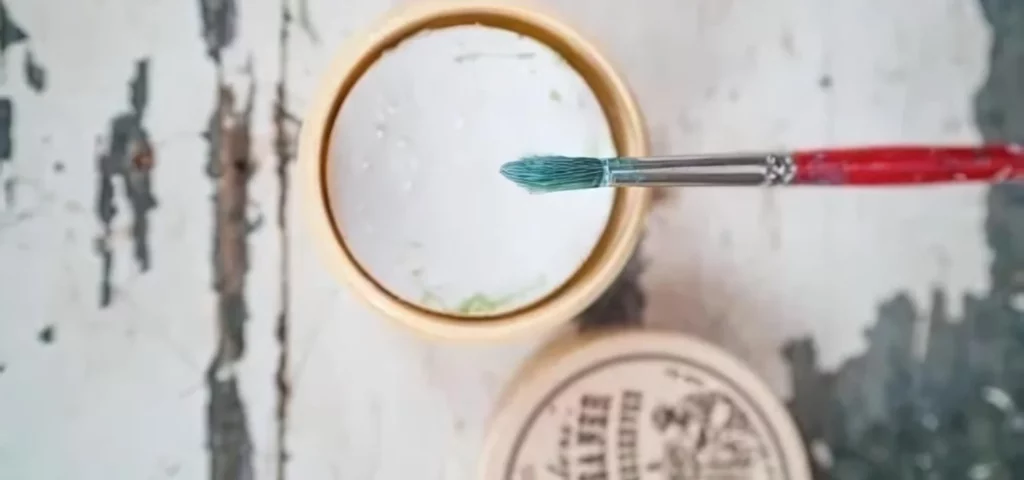 There is an open tub of Masters Brush Cleaner and Preserver in the center of the frame. The lid is to the left of the tub and there is a paint brush balancing over the tub. The tub is filled with a white paste. On a faux wooden surface.