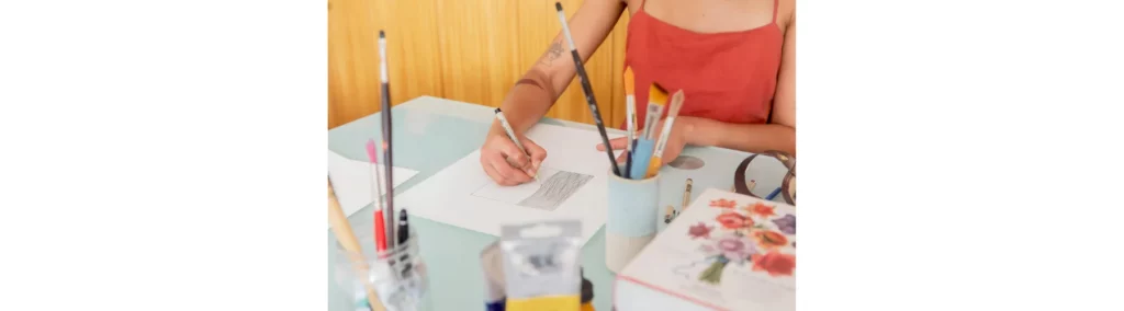 in the center of the image there is a woman in a red short sitting at a table facing toward us drawing on some paper. around her on the desk are various pens and brushes as well as papers and paintbrushes. behind her is a brown paneld wall. the image has a bright happy feel to it