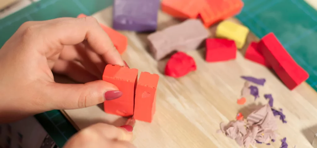 there are a few blocks of polymer clay on the brown wooden table surface. they are in red and brown colours with one purple square at the back left hand side of the image. on the left habd side front of the image is a pair of womans hands with red painted fingernails holding a block of red polymer clay in her hands about to cut and mould it. 