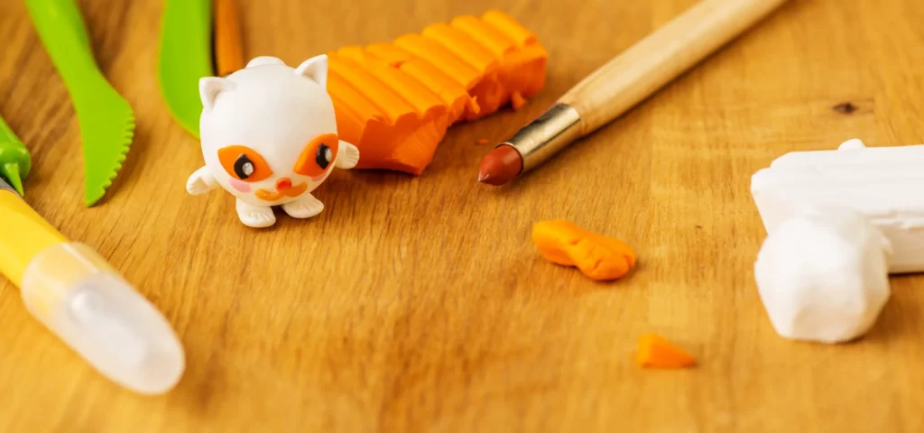 in the top middle of the image is the polymer clay block in an orange colur and a cut up square shape. infront of it is a tool that has been used and on the left of it is a figure that has been made using the clay. they are resting on a wooden table top. the image has a orange hue to it