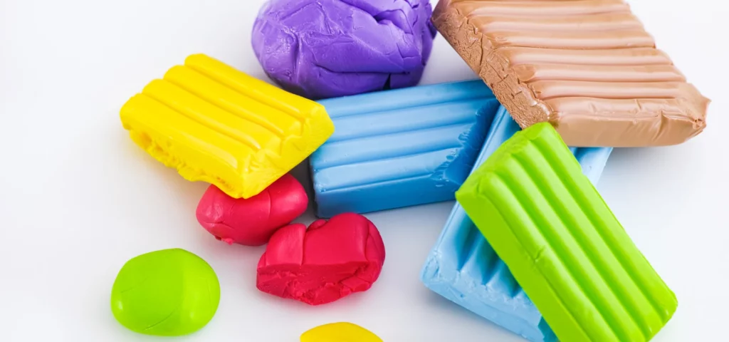 polymer clay is scattered all across the image in vibrant colours of blue, purple, yellow, red and gree, they are haphazardly stacked on top of eachother and are across the center of the image. they are on a white background. the blue, yellow and green are all in a square form and the purple and red are rolled into rough ball shapes