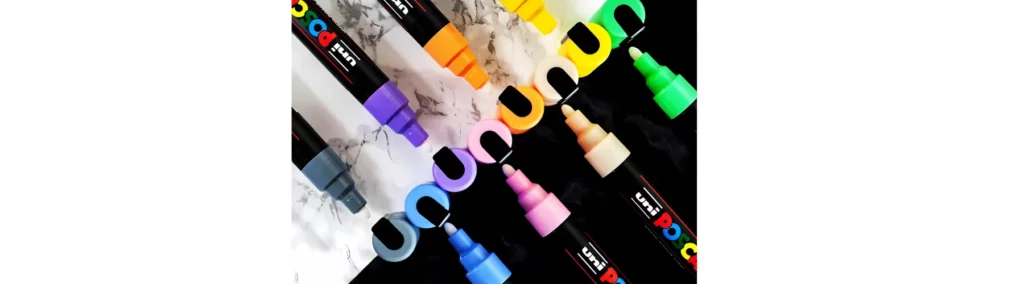 in the centre of the image are two rows of posca markers facing towards eachother. their caps are off and in the middle of the facing pens. they are a rainbow of colours. on half is on black and the other half is on white