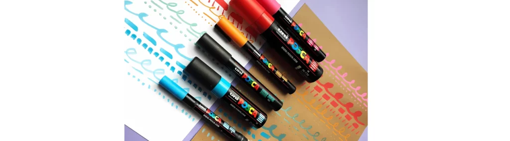 in the centre of the image there are a variety of posca pens on paper. one side of the paper is white the other is brown kraft paper. the posca has down squiggly lines on each type of paper in each of the colours that are in the centre of the image