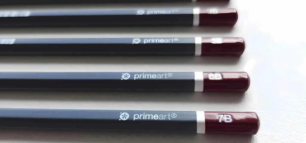 Prime Art Graphite Pencil Set. 9 pencils are laying diagonally across the center of the frame. The pencils have a maroon tip with the grade printed in white and a dark grey barrel. The maroon ends are facing the right hand side of the frame and the lead tips are facing towards the top left hand corner. They are on a light grey background with a canvas texture.