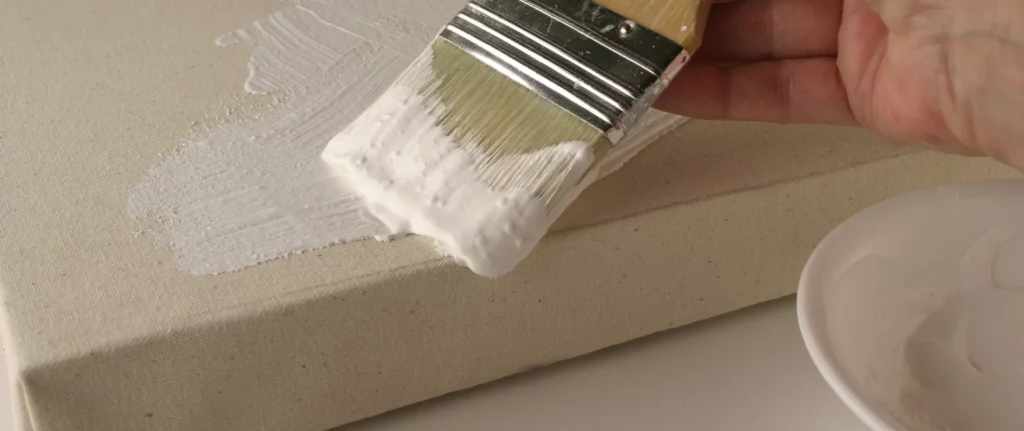 the image is of someone priming a canvas. the canvas is seen lying down and there is a varnish brush full of white gesso being used to apply the primer to the canvas. the primer is a brilliant white and the canvas is an off white ivory colour. there is a ceramic bowl to the right of the canvas. the persons hand can be seen at the top right of the image holding the paintbrush. you can see the canvas is resting on a white surface.