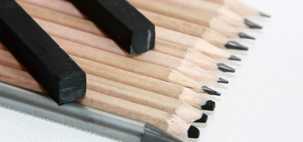 i the center of the image coming from the left hand side are brown wood cased pencils stacked tightly in a row and all facing the right hand side of the image. they all have black nibs. on top of them are two angled blocks of charcoal. next to them on the right is a graphite stick which is a grey colour. they are resting on a white surface.